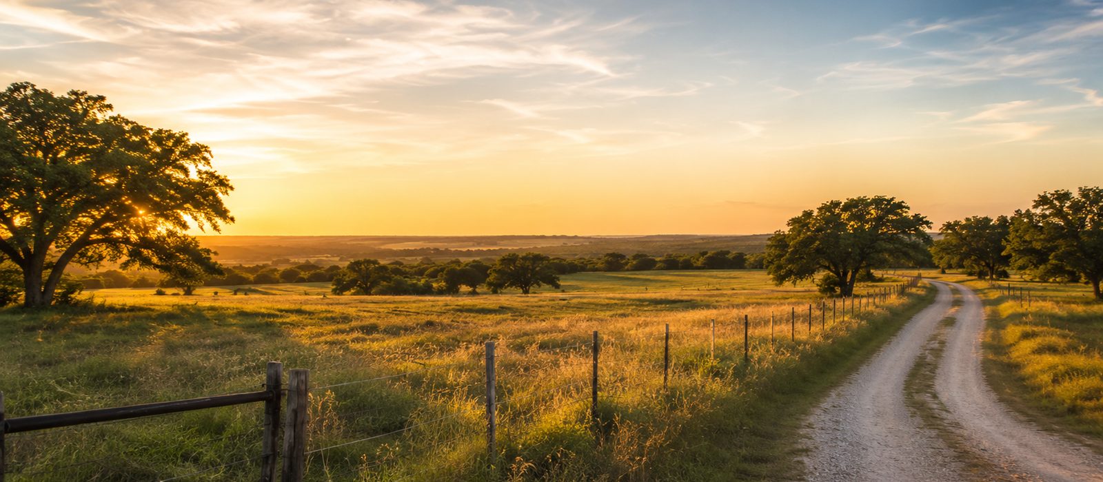 A Texas ranch entrance at golden hour, with a wooden fence and gate along a gravel road through native pasture under a wide sky.